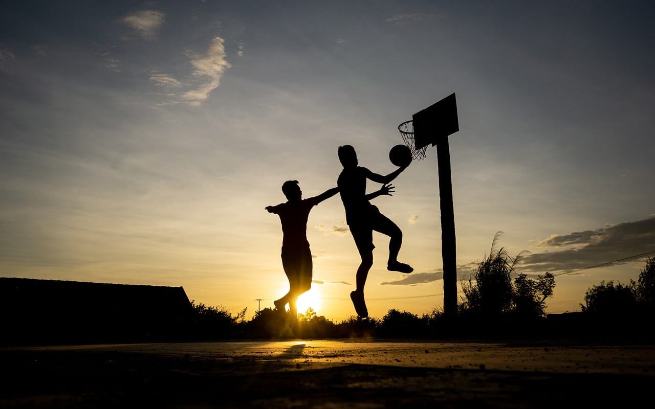 Deux jeunes joueurs en contre-jour au coucher du soleil, l’un montant au panier